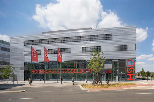 Dieses Bild zeigt das zentrale Hörsaalgebäude der Hochschule Düsseldorf mit einer Fassade aus Stahlblech und Glas. Im Erdgeschoss befinden sich die Hörsäle mit roten Stühlen. Im Obergeschoss gibt es Büroräume für die Beschäftigten der Hochschule.